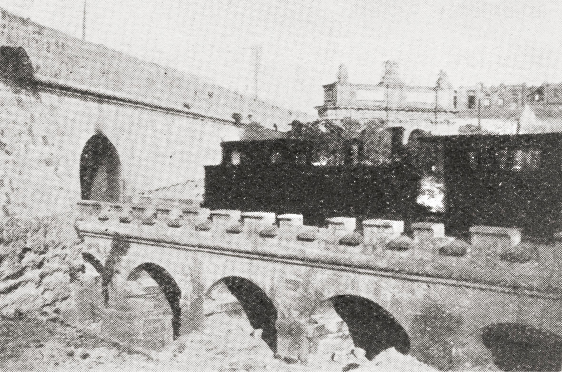 A great 1929 view of the fosse bray and Notre Dame curtain with Porte des Bombes visible beyond. Here, a Beyer Peacock engine returns a train to Valletta across the battlemented stone bridge before disappearing into the fortifications.  Whilst the original drop ditch below the tunnel mouth aligns with the angle of the defensive wall, the stone bridge piers are orthogonal with the alignment of the bridge. The half bridge arch on the left resulted from the clash of geometries.