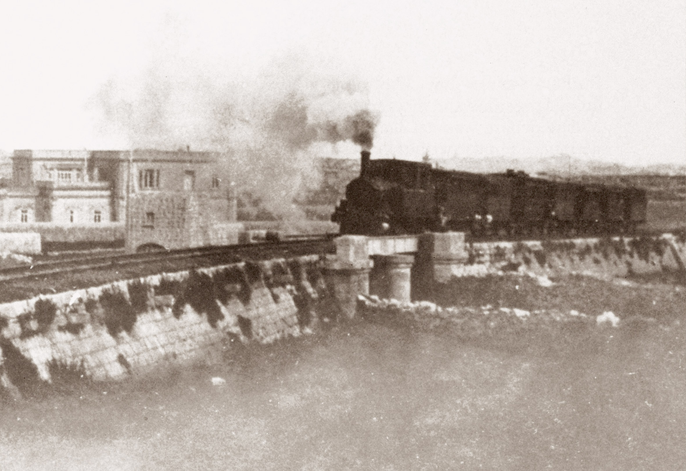 Heralded by a choking cough and clouds of smoke and steam, a heavy train labours uphill towards Attard, just about to cross the main road by way of the bridge. Long trains such as this were uneconomical to run beyond Birkirkara where there were fewer passengers.