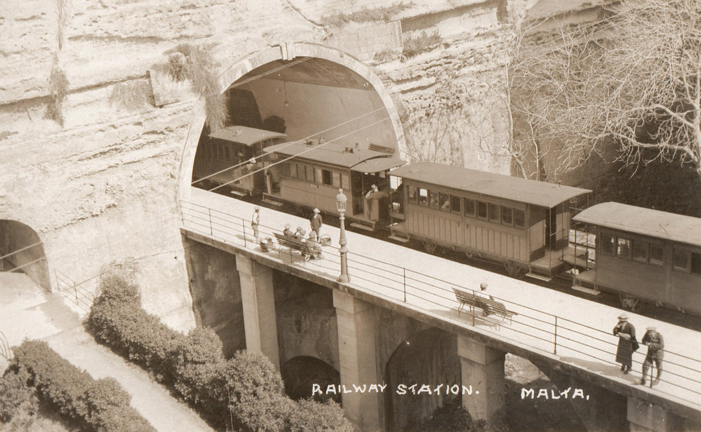 This early 20th century postcard view perfectly illustrates the later stone piers, added beside the existing bridge in about 1898 to support a wider station platform across the ditch. The arched buttresses supporting the main bridge arch against the wall of the ditch can be seen immediately below the station portal. The small tunnel opening and ramp on the far left were added in 1898 as public access to the city and station from the path through the main ditch. (Courtesy of Malta Study Circle)