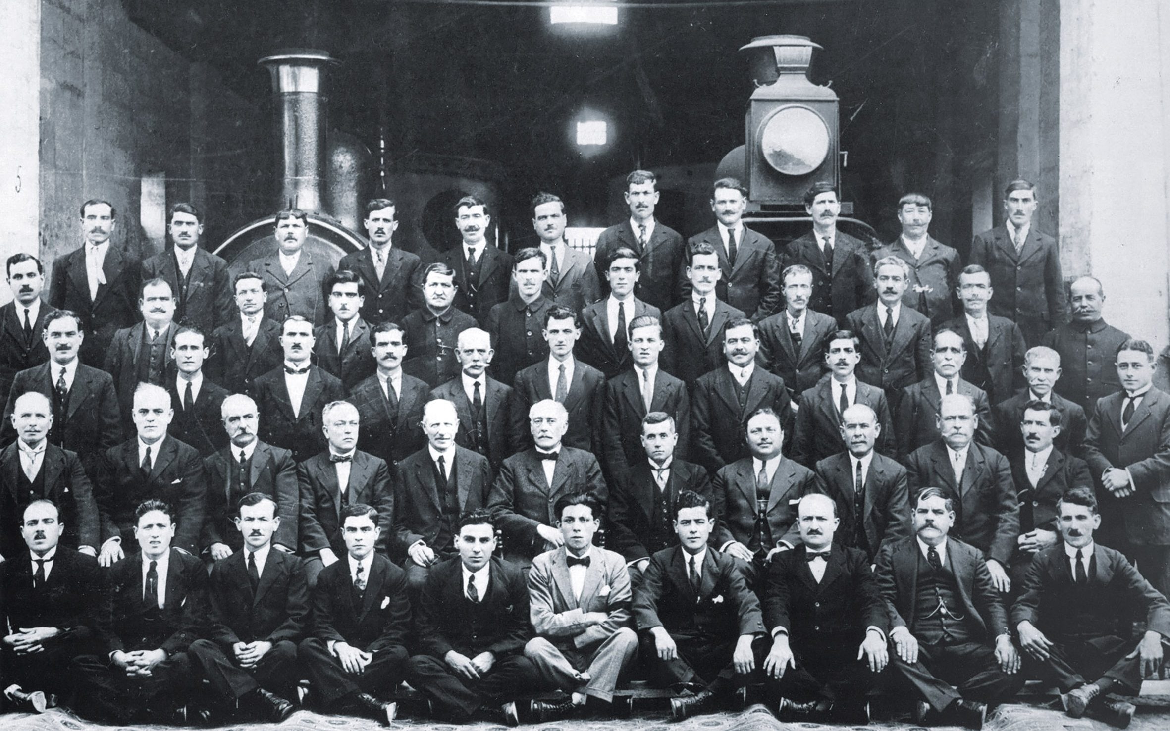 Railway and Technical School staff pose for a group photo in front of two of the railway's engines to commemorate the retirement of Nicola Buhagiar in 1924. Buhagiar is seated centrally in the second row from the front, recognisable in his bowtie. Sat to the left is Professor Nixon, of the University of Malta, who was Superintendent of the Technical school providing a practical engineering education at the workshops.