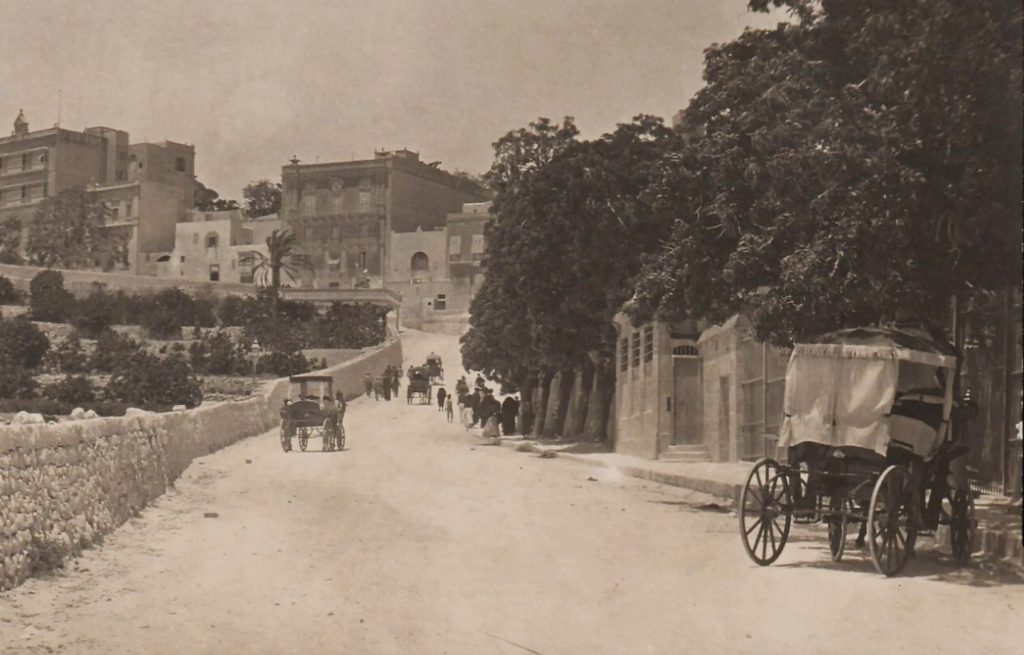 Looking up a road in Malta towards buildings on a hill beyond. Carriages and crowds head uphill from a train ot long arrived.
