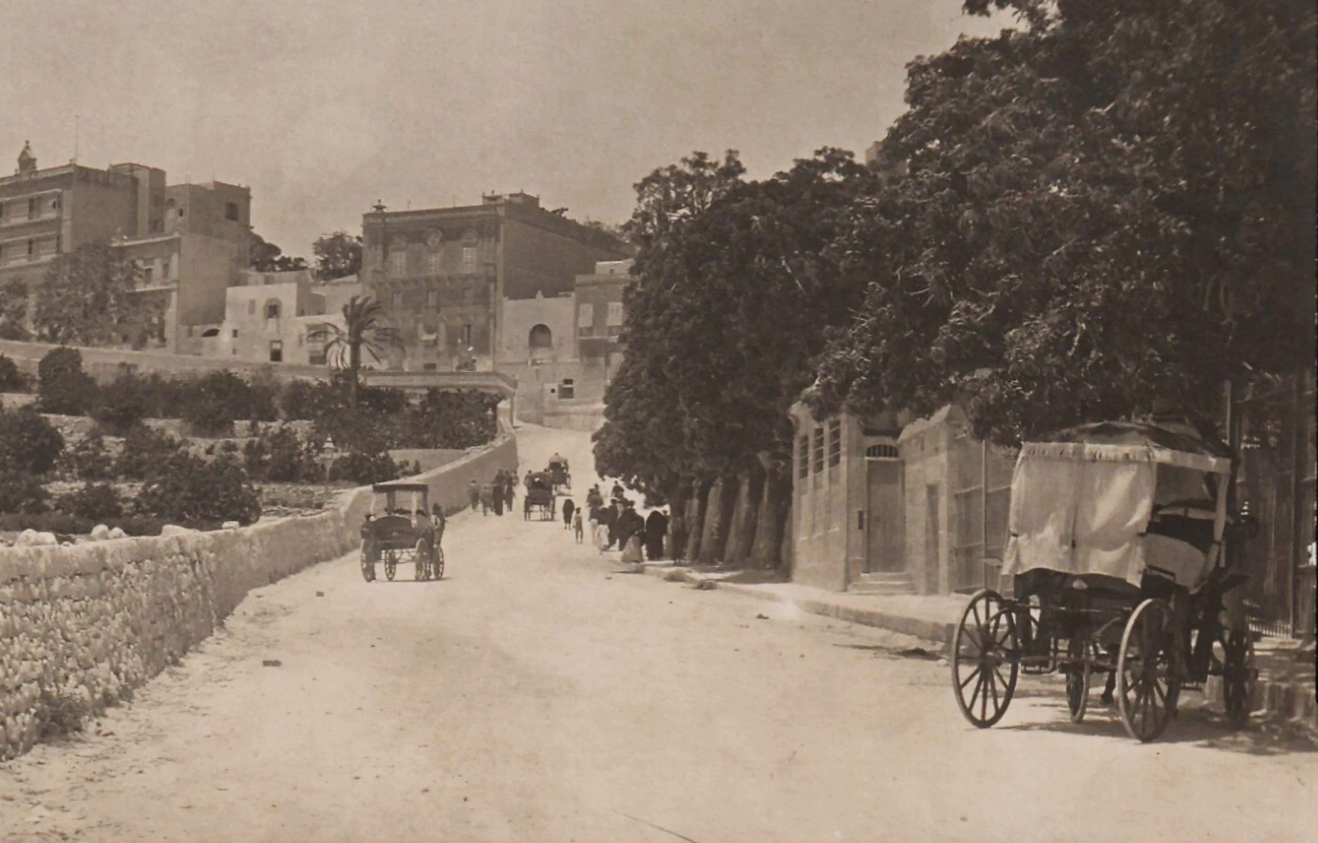 Looking up a road in Malta towards buildings on a hill beyond. Carriages and crowds head uphill from a train ot long arrived. 
